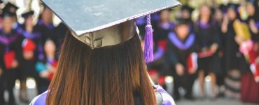 View of graduate's head from the back. They are wearing cap and gown, and other graduates similarly dressed in the background.